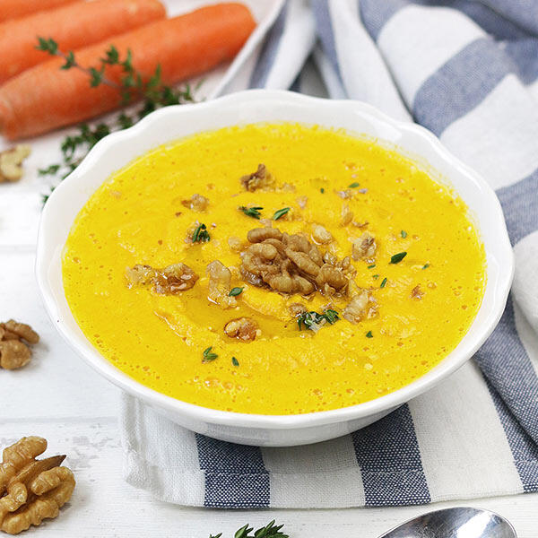 Rohköstliche Karottensuppe Eine Schale mit cremiger, gelber rohköstlicher Karottensuppe, garniert mit Walnusshälften und frischem Thymian. Im Hintergrund liegen zwei frische Karotten und ein blau-weiß gestreiftes Tuch.