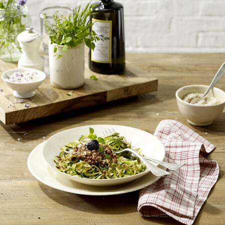 Eine Schale mit Zucchini-Pasta, garniert mit Paranuss-Parmesan und einem schwarzen Oliven-Topping. Daneben eine Gabel, ein rot-weiß kariertes Küchentuch, Kräuter in einem Topf und eine Flasche Olivenöl.