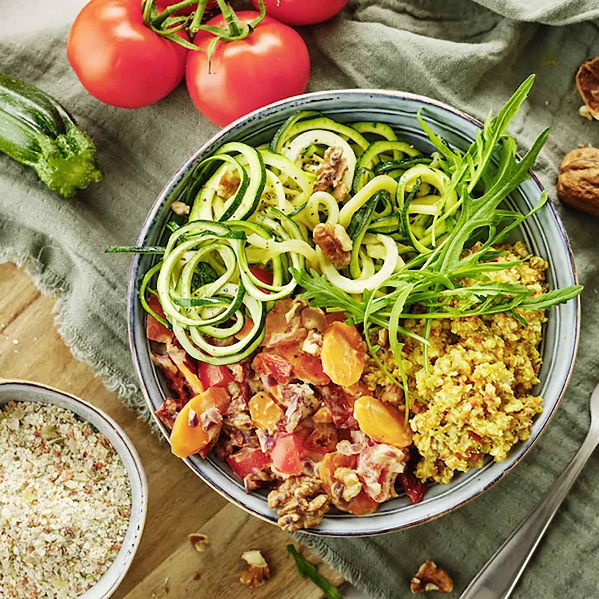 Zucchini-Nudel-Bowl mit Erdnusssoße Schüssel mit Zucchini-Nudeln, gewürfelten Tomaten und Möhren in Erdnusssoße, garniert mit grünen Blättern und Walnüssen, daneben frische Tomaten, Zucchini und eine Schale mit Körnern auf Stoff und Holz.