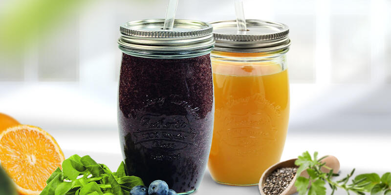 Ein dunkelvioletter und ein oranger Smoothie in Einmachgläsern mit Deckeln und Strohhalmen stehen nebeneinander auf einer weißen Fläche. Daneben liegen frische Heidelbeeren, Blätter, Orangenscheiben und Chiasamen.