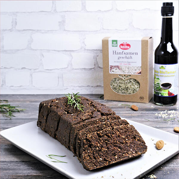 Pumpernickel, roh-vegan Dunkles, in Scheiben geschnittenes Pumpernickelbrot auf weißem Teller mit Rosmarinzweig. Im Hintergrund eine Packung geschälte Hanfsamen und eine Flasche Bio Tamari-Sojasauce.