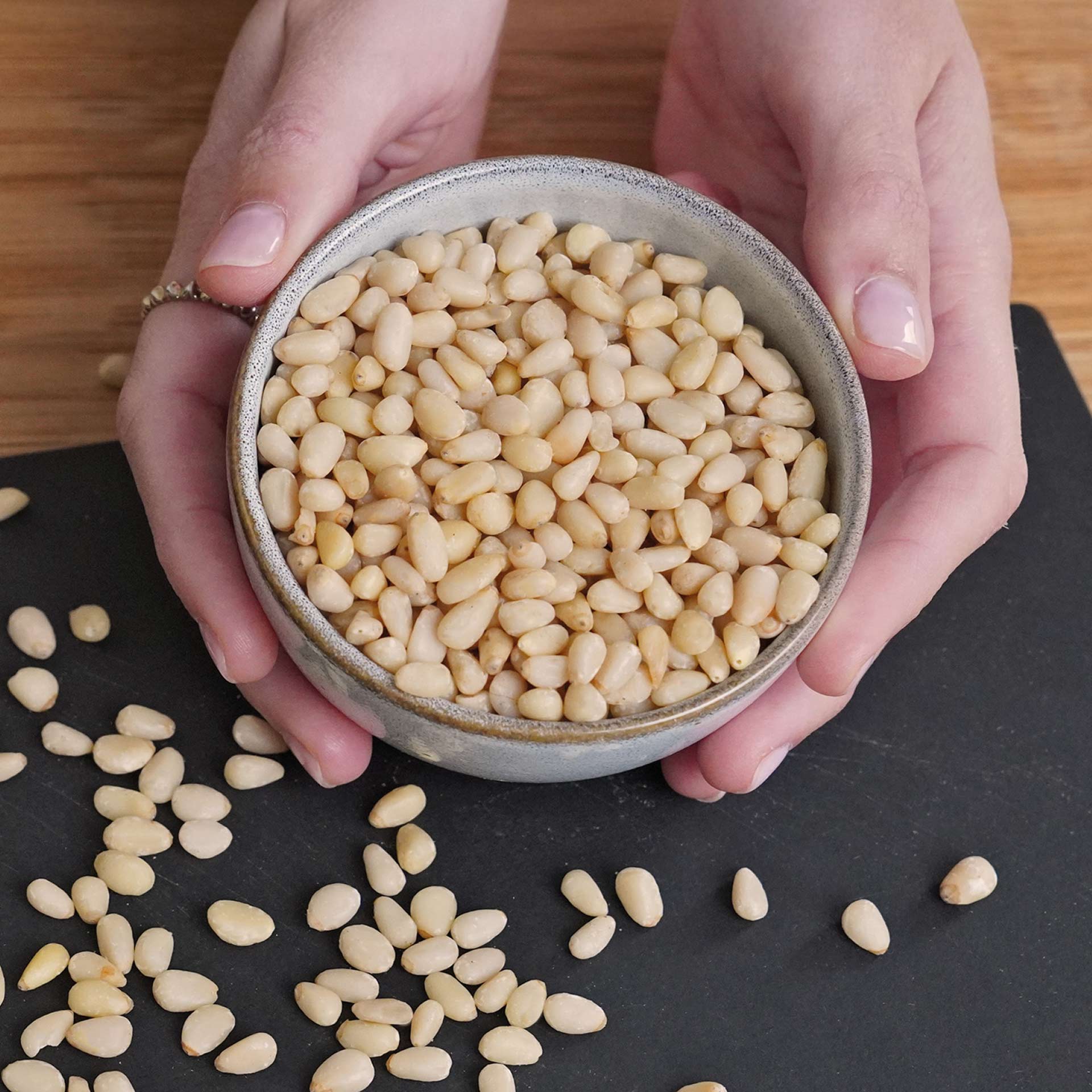 Zedernkerne, bio 100 g Hände halten eine graue, runde Schale, die mit vielen kleinen, hellen Pinienkernen gefüllt ist. Einige Pinienkerne liegen auf einer schwarzen Unterlage und einem Holzuntergrund verstreut.