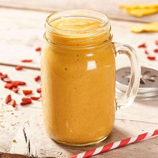 Mango Hanf Smoothie Ein Glas mit cremigem, orangefarbenem Mango-Hanf-Smoothie steht auf einem Holztisch. Daneben liegen verstreute rote Goji-Beeren und ein roter Strohhalm mit weißen Streifen.