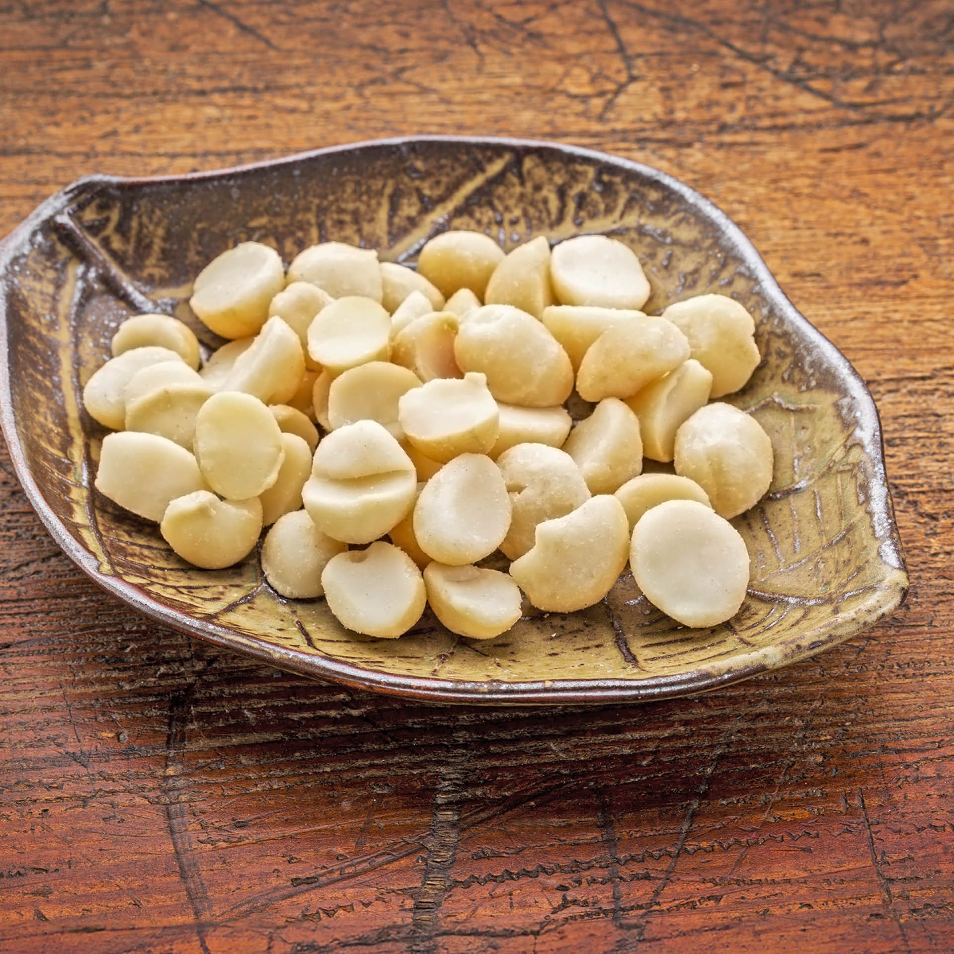 Eine braune, blattförmige Schale ist gefüllt mit vielen halben, hellen Macadamianüssen auf einem hölzernen Untergrund.