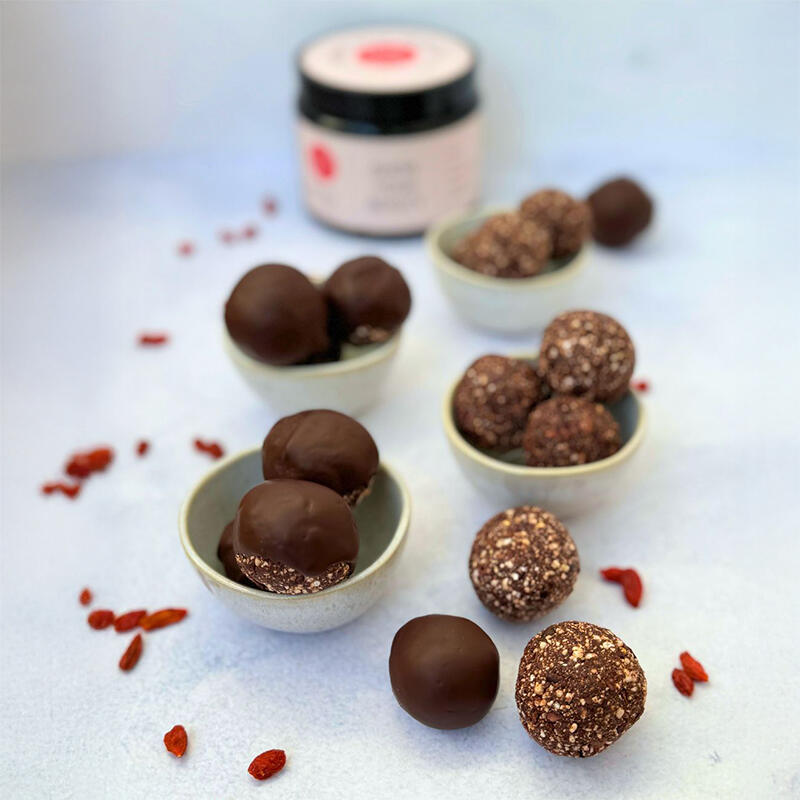 Energy-Boost Bällchen von Maria Sperling Drei kleine Schalen mit Energy-Boost-Bällchen, einige davon mit Schokoladenüberzug, auf weißem Untergrund verteilt. Im Hintergrund steht ein Glas mit weißem Etikett. Rote Beeren sind dekorativ verstreut.