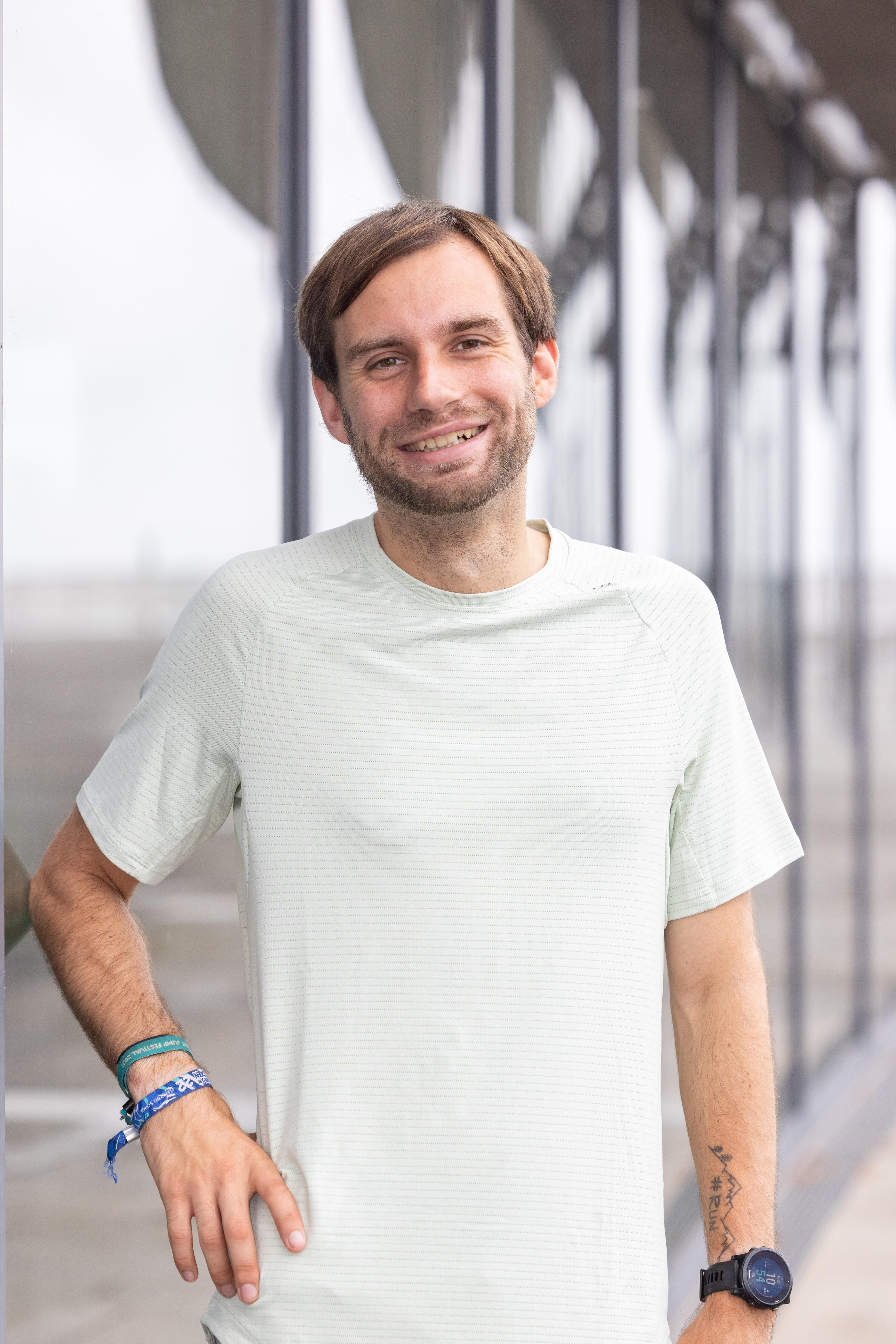 Jannick Lüttge steht draußen vor einer modernen Glasfassade und trägt ein helles T-Shirt. Er lächelt in die Kamera und hat eine Armbanduhr sowie mehrere Armbänder am Handgelenk.