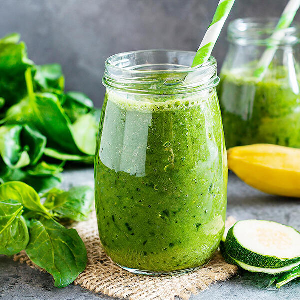 Green Power Smoothie von @vegan.food.investigation Ein Glas gefüllt mit grünem Smoothie steht auf einem Stoffunterlage. Im Glas steckt ein grüner Strohhalm. Daneben liegen Babyspinatblätter, eine halbe Zucchini und eine gelbe Banane. Im Hintergrund steht ein weiteres Glas mit grünem Smoothie.