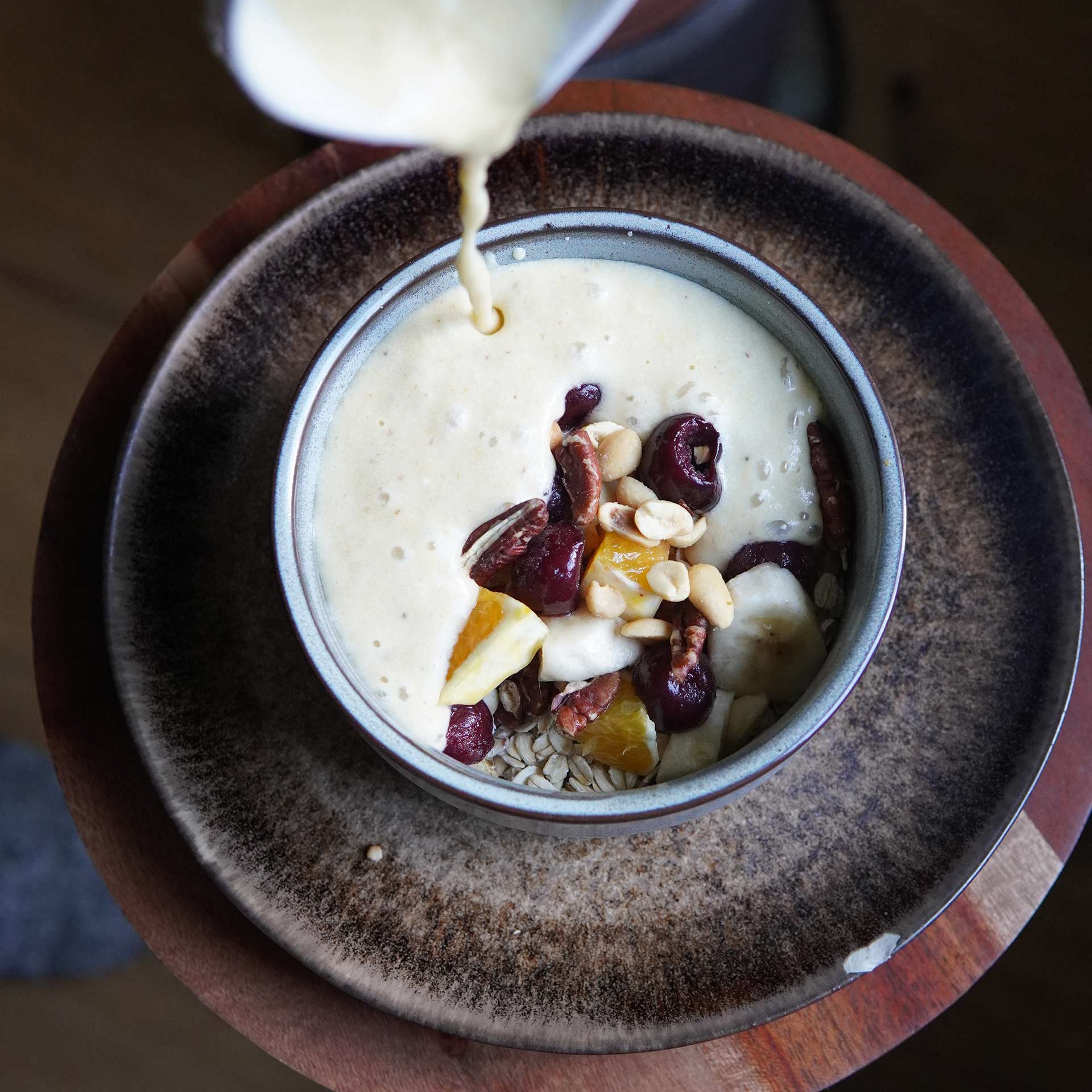 Cheesecake Porridge Frisch zubereiteter, cremiger Porridge mit Kirschen, Orangen, Bananenscheiben, Haferflocken, Pekannüssen und Erdnüssen in einer Schale. Die cremige Masse wird gerade darüber gegossen.