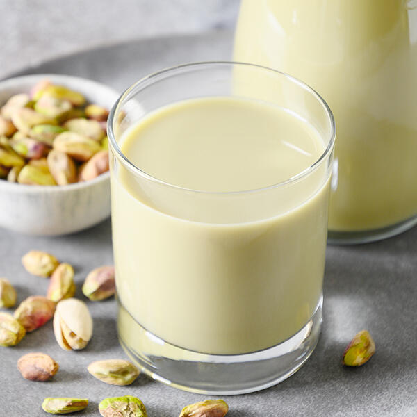 Rohe Pistazienmilch von Peter Dreverhoff Ein Glas rohe Pistazienmilch steht im Vordergrund. Im Hintergrund ist eine Karaffe mit der Milch und eine Schale mit rohen Pistazien zu sehen. Rohe Pistazien liegen auf der grauen Oberfläche verstreut.