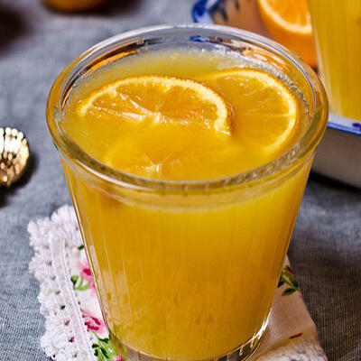 Orangencreme Ein Glas mit gelber Orangencreme steht auf einer bestickten Stoffunterlage. Zwei Orangenscheiben schwimmen oben auf der cremigen Masse.