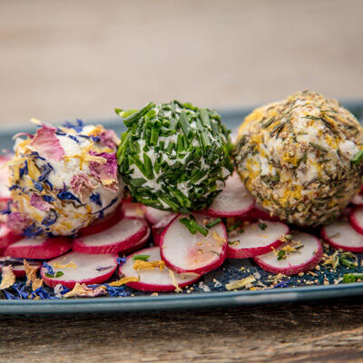 Drei Kugeln Mandel-Ricotta-Käse sind mit essbaren Blüten, Schnittlauch und Gewürzen bedeckt. Sie liegen auf einem Teller mit dünnen Radieschenscheiben.