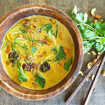 Rohkost-Rezept: Raw Miso Ramen von Boris Lauser Eine Schale Raw Miso Ramen in einer Holzschale mit grünen Kräutern, Pilzen und Cashewkernen. Daneben liegen frischer Koriander, Cashewkerne und Holzstäbchen.