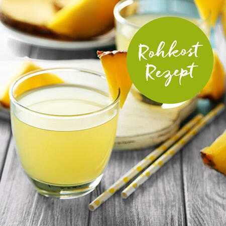 Zwei Gläser mit hellgelbem Saft stehen auf einem Holztisch. Eins der Gläser ist mit einem Ananasstück garniert. Daneben liegen zwei weiße Trinkhalme mit gelben Punkten. Auf dem Bild ist ein grüner Kreis mit dem Text Rohkost Rezept.