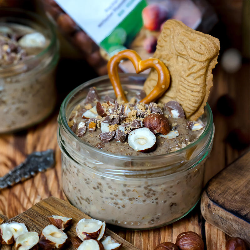 Weihnachtliche Overnight Oats Ein Glas mit Overnight Oats, garniert mit gehackten Haselnüssen, Schokoladenstücken, einem Brezelherz und einem Lebkuchenplätzchen in Form einer Eule.