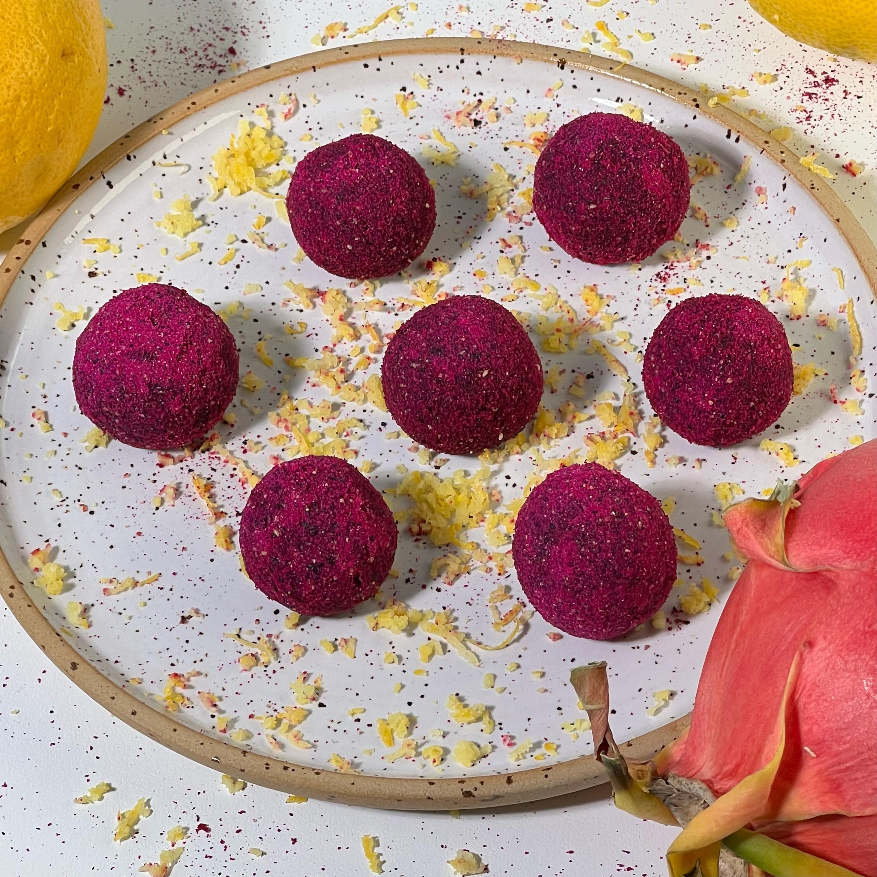 Pink Power Beauty Balls Sieben kugelförmige, dunkelrote vegane Süßigkeiten liegen auf einem weißen Teller mit braunem Rand. Der Teller ist mit Zitronenschale bestreut. Rechts unten und links außen sind eine Drachenfrucht und eine Zitrone zu sehen.