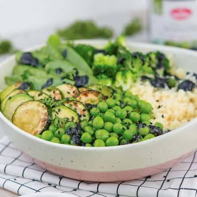 Buddha-Bowl mit schwarzem Sesammus-Dressing von Deutschland is(s)t vegan Buddha-Bowl mit Zucchinischeiben, grünen Erbsen, Brokkoliröschen, Hirse und grünem Blattgemüse, garniert mit schwarzem Sesam in einer weißen Schale auf einem karierten Tuch.