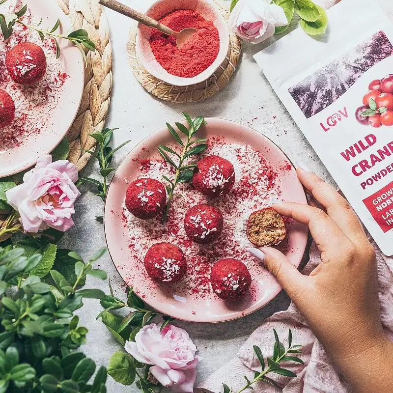 Cranberry-Erdnuss Energy-Balls Fünf rote Cranberry-Erdnuss Energy-Balls mit Kokosraspeln auf einem Teller liegen, daneben hält eine Hand ein halbiertes Energy-Ball. Daneben steht ein Beutel mit der Aufschrift LOOV WILD CRANBERRY POWDER.