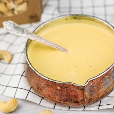 Ein kupferfarbener Topf mit cremiger, hellgelber Cashew-Creme steht auf einem karierten, weißen Tuch. Ein silberner Löffel steckt in der Creme. Cashewkerne liegen daneben.
