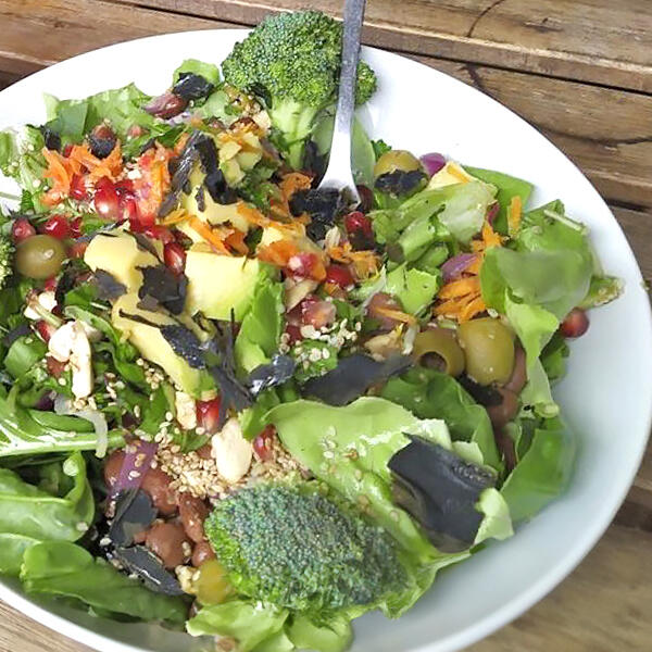 Protein Powersalat mit Sprossen und Keimlingen Weisser Teller mit einem bunten Powersalat aus Blattsalaten, Broccoli, grünen Oliven, roten Bohnen, geraspelten Karotten, Granatapfelkernen und schwarzem Algen-Topping. Ein Löffel steckt im Salat.
