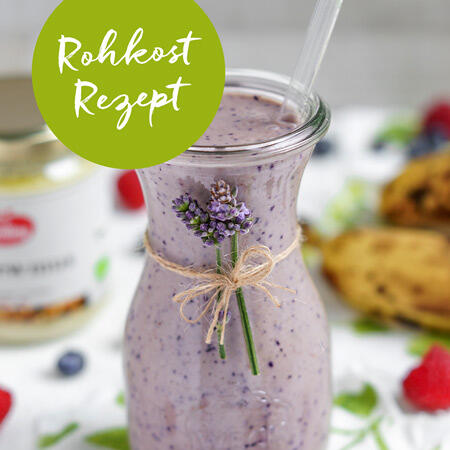 Glasflasche mit lila Smoothie, verziert mit einem Strauß Lavendel, im Hintergrund unscharfe Früchte und Zutaten. Grüner Kreis mit weißem Text Rohkost Rezept oben links.