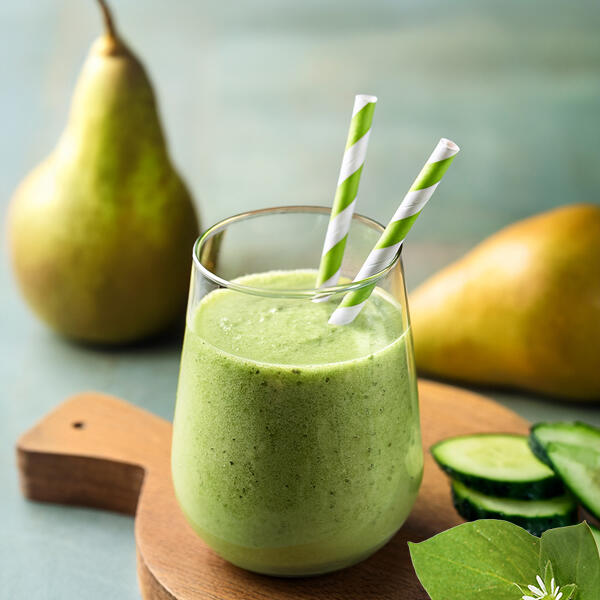 Grüner Frischekick Ein Glas grüner Smoothie mit zwei grünen, gestreiften Papierstrohhalmen steht auf einem Holzbrett. Im Hintergrund sind zwei ganze Birnen und einige Gurkenscheiben zu sehen.