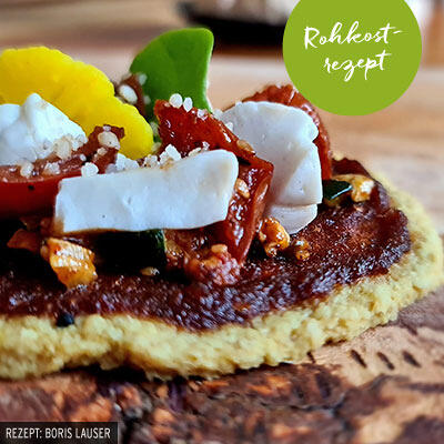 Orientalische Blumenkohl Pitas Pita mit einer roten Paste, belegt mit weißen Stücken, getrockneten Tomaten, grünen Blättern und einem gelben essbaren Blütenblatt. Oben rechts ein grüner Kreis mit dem weißen Text Rohkostrezept.