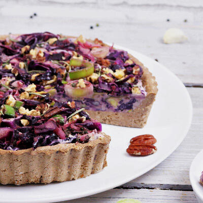 Rotkohl-Tarte, roh-vegan Rotkohltarte mit nussigem Boden und cremiger Cashew-Sahnecreme, belegt mit Rotkohl, Lauch, Apfelstücken und Nüssen, serviert auf weißem Teller auf hellem Holztisch.