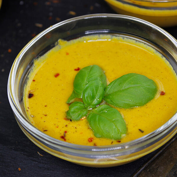 Weihnachtliche Rohkost-Suppe mit Cashews und Gojibeeren Gelbe Rohkost-Suppe in einer Glasschale, garniert mit drei frischen Basilikumblättern und roten Chiliflocken, steht auf einer dunklen Oberfläche.
