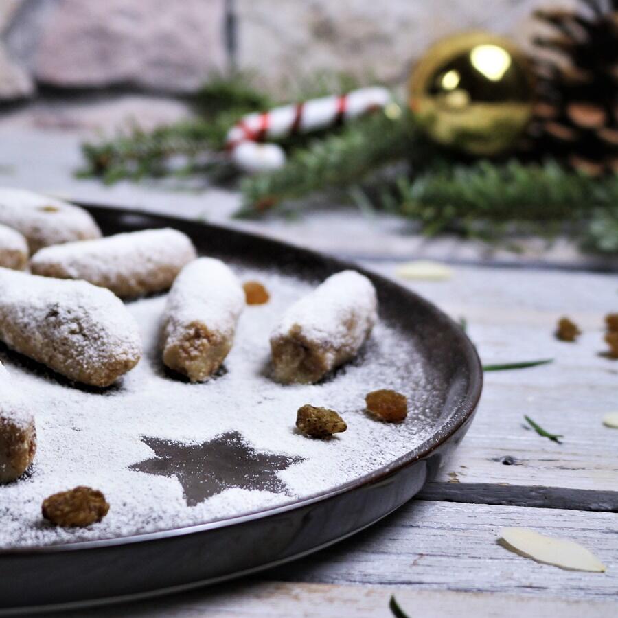 Raw Stollenkonfekt, roh-vegan Rohe Stollenkonfekt-Plätzchen liegen auf einem Teller, bestäubt mit Puderzucker, der einen Stern in der Mitte freilässt. Im Hintergrund sind Weihnachtsdekorationen und eine Zuckerstange unscharf zu sehen.