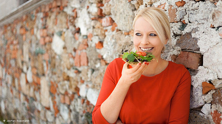 Michaela Russmann lehnt lächelnd an einer alten Ziegelmauer und hält ein mit frischem Gemüse belegtes Brotstück zum Mund.