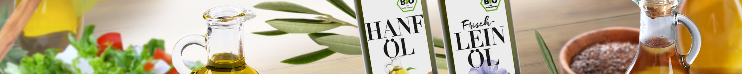 Zwei Flaschen mit Öl stehen auf einem hellen Holztisch. Die linke Flasche ist mit der Aufschrift HANF ÖL und die rechte mit Frisch LEIN ÖL beschriftet. Daneben befinden sich kleine Gläser mit Öl und eine Schale mit Samen.