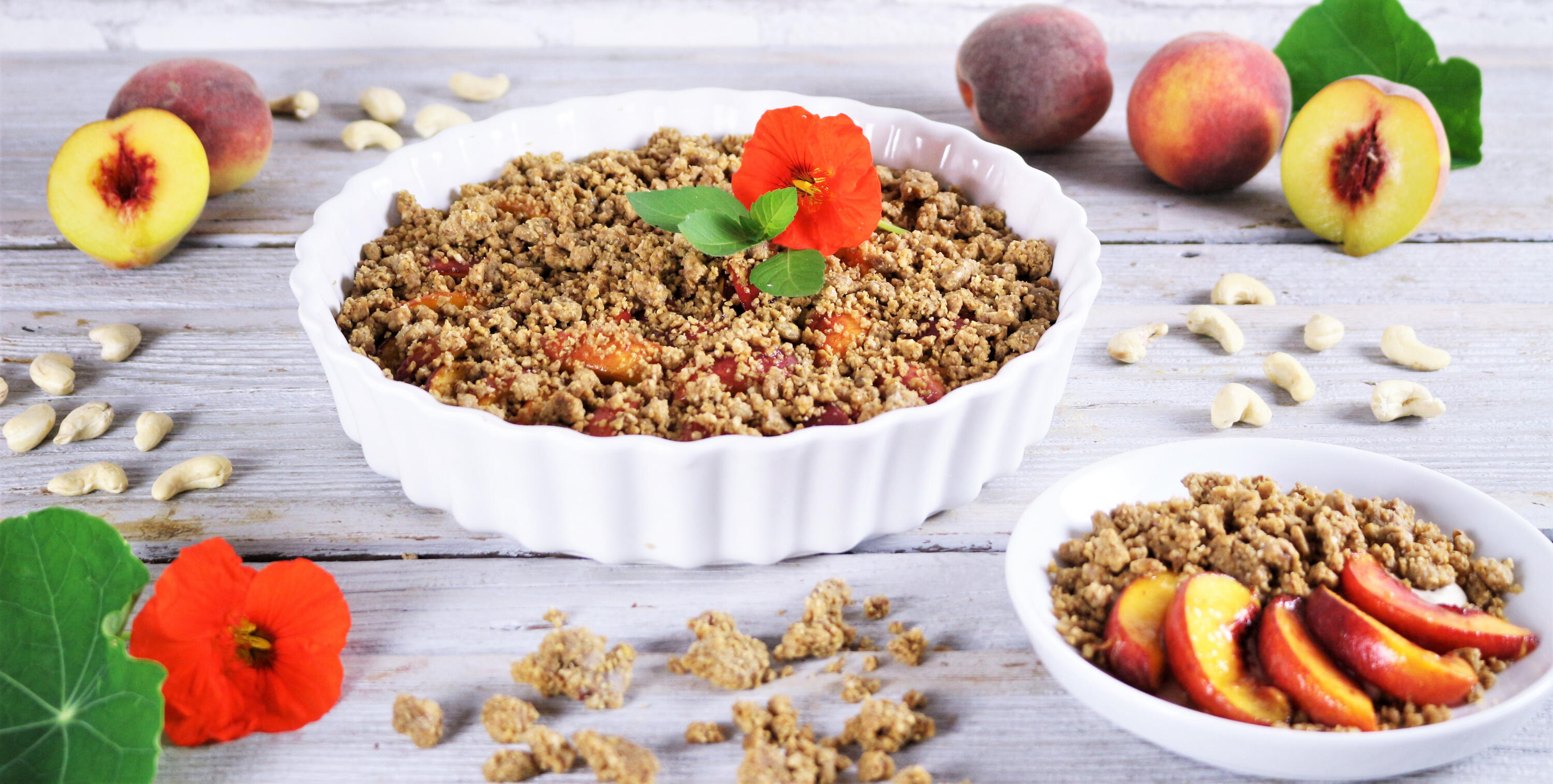 Weiße Keramikform mit Rohkost-Pfirsichcrumble, garniert mit einer roten Blüte und grünen Blättern. Dazu eine kleine weiße Schale mit Pfirsichscheiben und Streuseln. Frische Pfirsiche und Cashewkerne liegen auf einem hellen Holztisch.