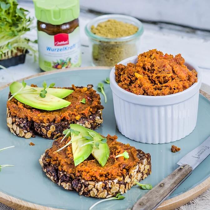 Feiner WurzelKraft Aufstrich mit Tomaten & Oliven Zwei Scheiben Vollkornbrot mit Nusskuchenaufstrich und Avocadoscheiben liegen auf einem blauen Teller. Daneben steht eine weiße Schale mit mehr Aufstrich und ein Messer.