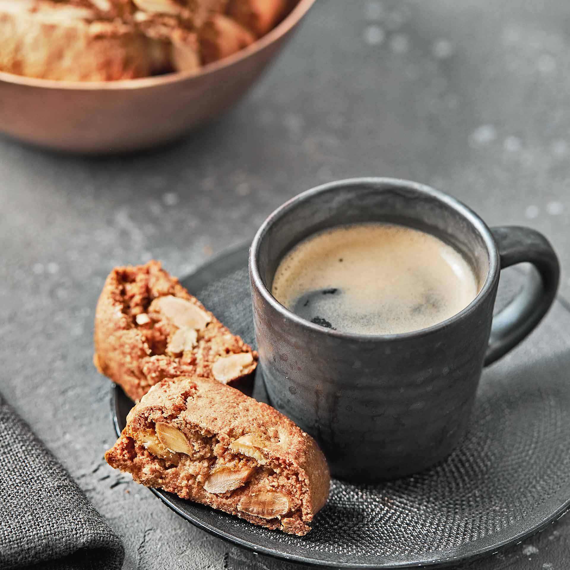 Vegane Cantuccini Eine schwarze Tasse mit Espresso steht auf einer schwarzen Untertasse. Daneben liegen zwei vegane Cantuccini mit sichtbaren Mandelstücken. Im Hintergrund ist leicht unscharf eine Schale mit weiteren Cantuccini.