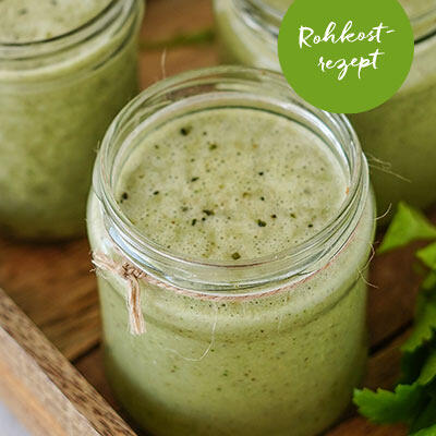 Glas mit grünem, schaumigem Saft aus Frankfurter Grüner Sauce steht auf Holztablett. Grüner Kreis mit weißem Text Rohkostrezept befindet sich am oberen Rand.