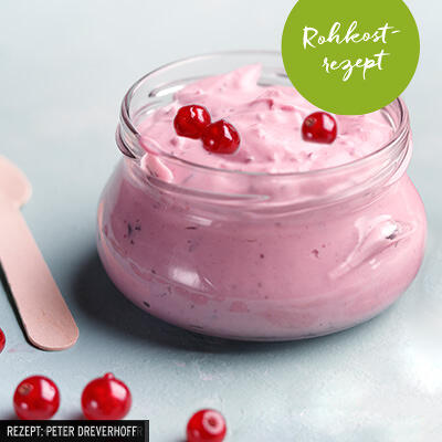 Schüssel mit rosa rohem, veganem Beerendessert, oben mit vier roten Beeren dekoriert. Grüner Kreis mit weißem Text Rohkostrezept auf dem Glas angebracht. Mehrere einzelne rote Beeren liegen neben der Schüssel.