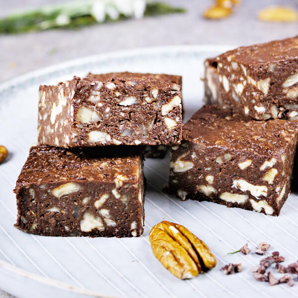 Crunchige Pekannuss-Schoko Schnitten, roh-vegan Vier rohe vegane Pekannuss-Schoko Schnitten liegen auf einem weißen Teller. Die Schnitten haben eine braune, schokoladige Farbe mit sichtbaren Pekannuss-Stückchen. Eine Pekannuss liegt vor den Schnitten.