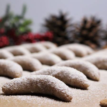 Mehrere rohe Vanillekipferl liegen auf einer Holzfläche und sind mit Puderzucker bestreut. Im Hintergrund sind unscharf Tannenzapfen und rote Beeren zu sehen.