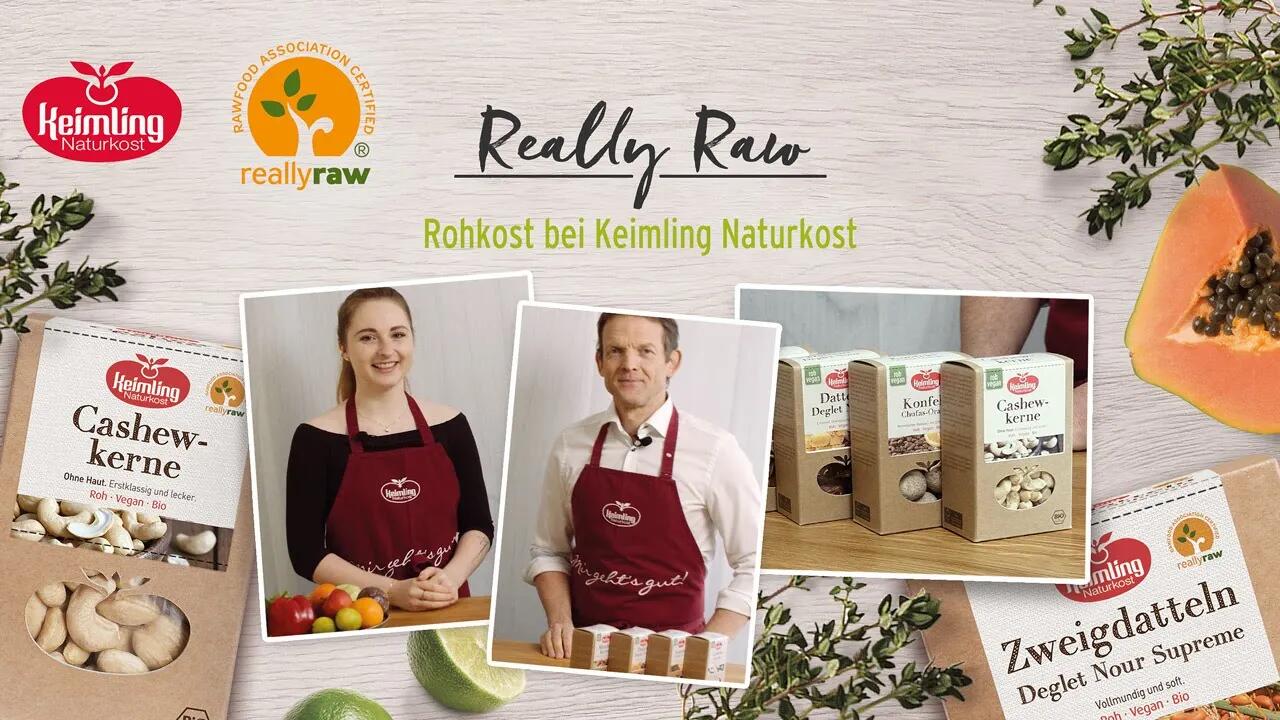 Zwei Personen in roten Keimling Naturkost-Schürzen präsentieren verschiedene Rohkost-Produkte wie Cashewkerne, Konfekt und Zweigdatteln auf einem hellen Holztisch.