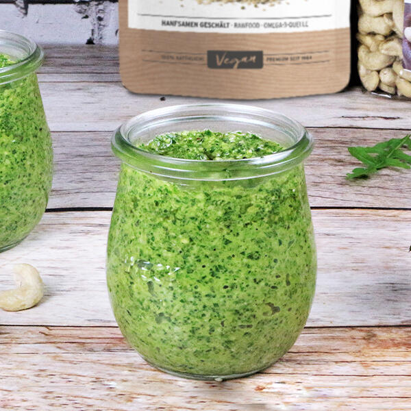 Rucola-Hanf-Petersilien Pesto, roh-vegan  Ein kleines Glas mit grünem Rucola-Hanf-Petersilien-Pesto steht auf einem Holzbrett. Im Hintergrund sind ein Behälter mit Hanfsamen und ein Paket mit der Aufschrift Vegan zu sehen.