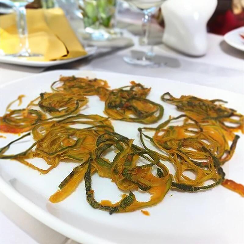Knusprige Rohkost Zucchini-Spaghetti Mehrere knusprige Zucchiniringe liegen auf einem weißen Teller. Im Hintergrund sind ein Glas und eine Serviette auf einem gedeckten Tisch zu sehen.