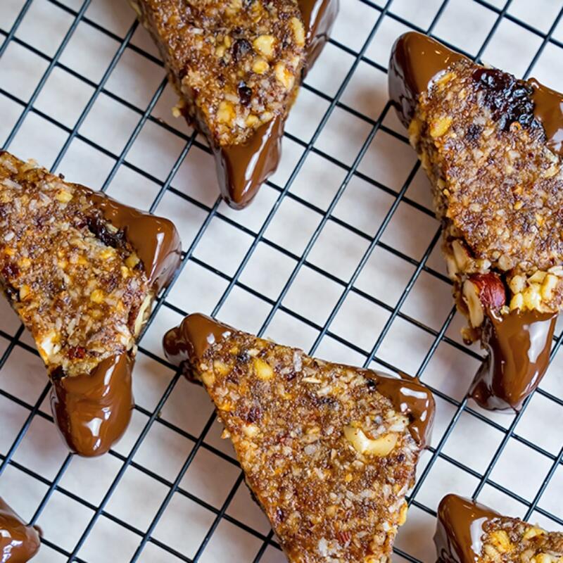 Vegane Nussecken mit Schokolade Dreieckige vegane Nussecken mit Mandeln, Walnüssen und Cranberries liegen auf einem schwarzen Gitterrost. Die Ecken sind in geschmolzene Zartbitterschokolade getaucht.
