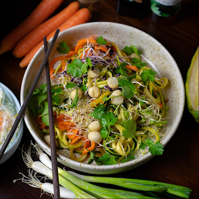 Veganer Pad-Thai-Salat mit Zucchini und Macadamia Schüssel mit veganem Pad-Thai-Salat aus Zucchini- und Karottenspiralen, frischen Sprossen, Macadamianüssen und Korianderblättern daneben liegen Frühlingszwiebeln, Karotten und Holz-Stäbchen.