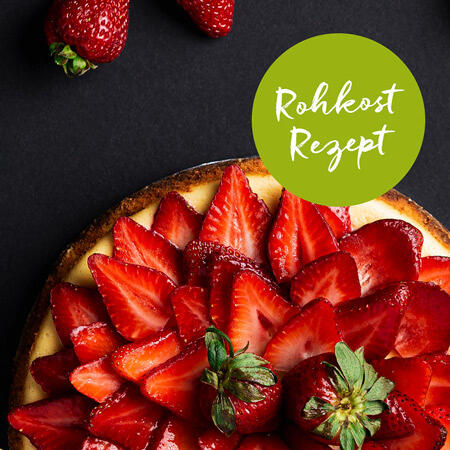 Eine rohköstliche Erdbeertorte mit frischen, aufgeschnittenen und ganzen Erdbeeren auf einer cremigen Creme. Auf der oberen rechten Seite steht in einem grünen Kreis der Text Rohkost Rezept.