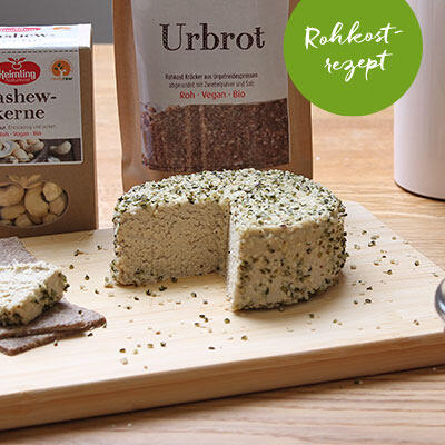 Runder, roh-veganer Käse mit Hanfsamen bestreut steht auf einem Holzbrett. Im Hintergrund sind eine Packung Urbrot und eine Packung Cashewkerne zu sehen. Ein grüner Kreis mit der Aufschrift Rohkostrezept ist eingeblendet.