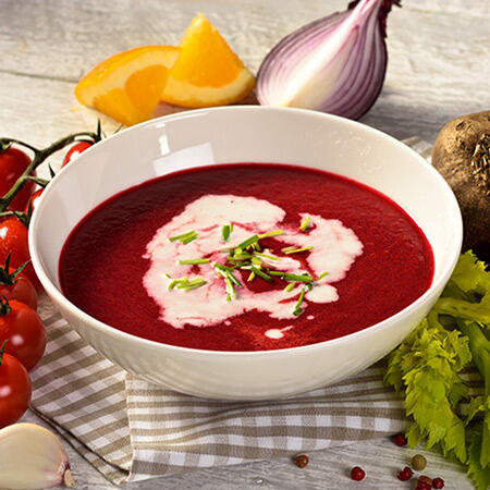 Rohkost Rote Bete Suppe Eine Schale mit roter Rote-Bete-Suppe steht auf einem karierten Tuch. Die Suppe ist mit weißer Mandelcreme und grünen Kräutern garniert. Im Hintergrund liegen eine Zwiebel, Orangenscheiben, Tomaten und Sellerie.