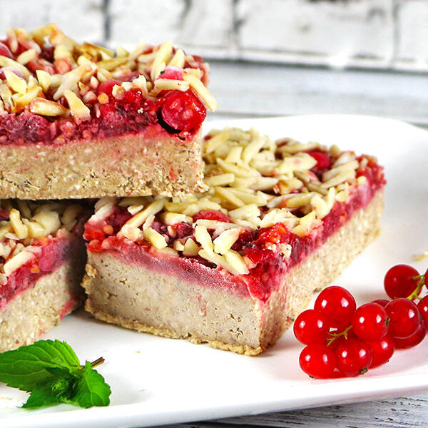 Leichter Johannisbeer-Mandelkuchen, roh-vegan Drei Stücke eines rohen, veganen Johannisbeer-Mandelkuchens mit einer hellen Teigschicht, einer roten Johannisbeerschicht und Mandelstiften als Topping liegen auf einem weißen Teller. Daneben liegen frische rote Johannisbeeren und ein Minzblatt.