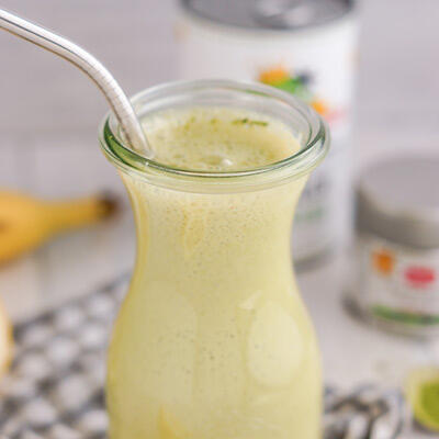 Ein Glas mit cremigem, hellgrünem Matcha-Kokos-Smoothie mit einem Metallstrohhalm steht auf einem karierten Tuch. Im Hintergrund sind unscharf Bananen und Dosen zu sehen.