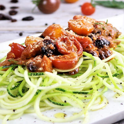 Rohkost-Rezept: Tomaten-Koriander-Cranberry Marinara mit Zucchinipasta Zucchininudeln liegen auf einem weißen Teller. Darauf befindet sich eine marinierte Tomaten-Koriander-Cranberry-Sauce mit halben Kirschtomaten und Cranberries. Das Gericht ist roh-vegan und low carb.