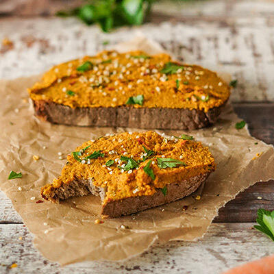 Kürbis-Möhren-Aufstrich Zwei Scheiben Brot sind mit einem orangenen Kürbis-Möhren-Aufstrich bestrichen. Der Aufstrich ist mit frischen Kräutern und weißen Sesamsamen garniert. Eine Scheibe hat einen Biss.
